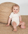 A baby in a white LW655 by Larkwood sits on a woven rug, leaning against a large, round, woven basket, and smiling.