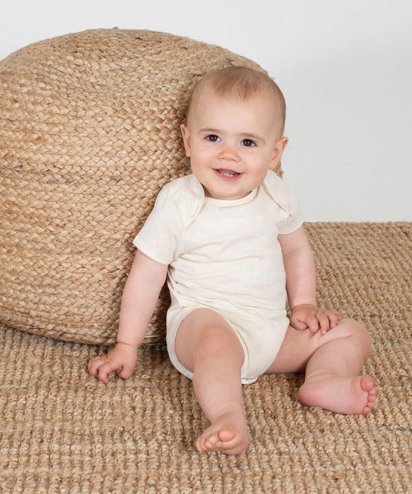 A baby in a white LW655 by Larkwood sits on a woven rug, leaning against a large, round, woven basket, and smiling.