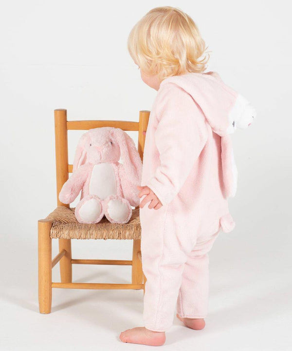 A toddler in a pink Larkwood LW73T with bunny ears stands facing a wooden chair with a pink plush bunny toy on it.