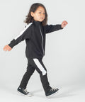 A child with medium-length hair walks forward wearing a Larkwood LW830 black tracksuit with white stripes and black sneakers against a plain background.