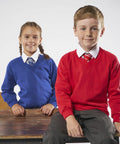 Two children sitting on a bench, dressed in school uniforms; the girl wears a blue MD02B Maddins sweater and tie, while the boy wears a red MD02B Maddins sweater and tie. Both are smiling and looking at the camera.