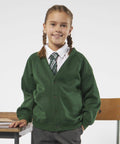 A young girl with brown hair in braids, wearing a green cardigan, white shirt, striped tie, and gray pants, stands next to a wooden chair and desk. She is smiling and facing the camera. The wooden chair and desk are from the Maddins MD04B collection.