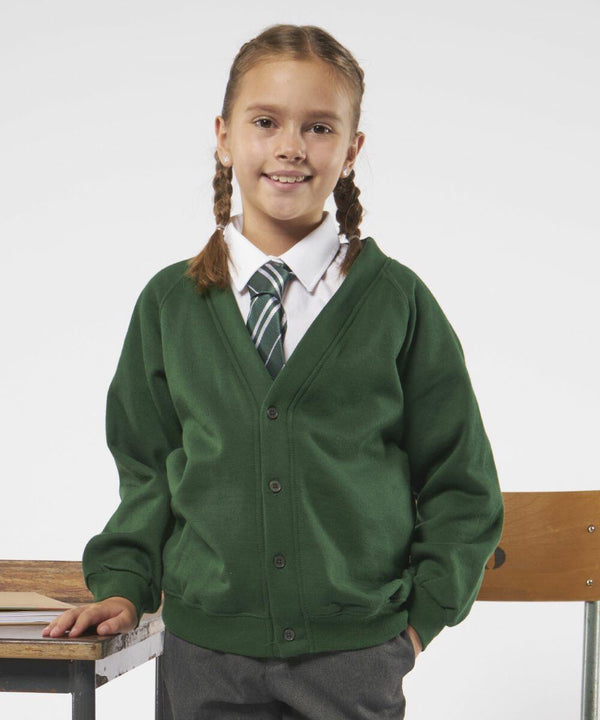 A young girl with brown hair in braids, wearing a green cardigan, white shirt, striped tie, and gray pants, stands next to a wooden chair and desk. She is smiling and facing the camera. The wooden chair and desk are from the Maddins MD04B collection.