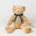 The MM001 teddy bear from Mumbles, featuring a beige fur and a blue plaid ribbon, sits upright against a plain white background.