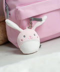 A Mumbles Character keyring, a small bunny plush with white and pink fur, is attached to a light pink rucksack resting on a light surface.