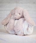 A soft, plush bunny toy with long ears and a round body wrapped in a white blanket, placed on a light-colored surface, MM034 by Mumbles.