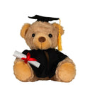 A soft toy brown teddy bear dressed in a black graduation cap with yellow tassel, black gown, and holding a rolled-up diploma tied with a red ribbon.