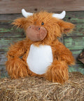 A Mumbles MM060 resembling a highland cow with orange fur, white horns, and a white belly, sitting on a bed of straw against a rustic wooden background.