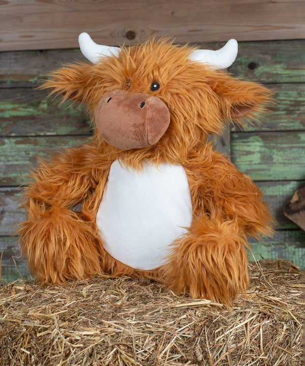 A Mumbles MM060 resembling a highland cow with orange fur, white horns, and a white belly, sitting on a bed of straw against a rustic wooden background.