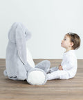 A toddler in a white outfit sits on the floor facing a large grey stuffed bunny, the MM550 by Mumbles.