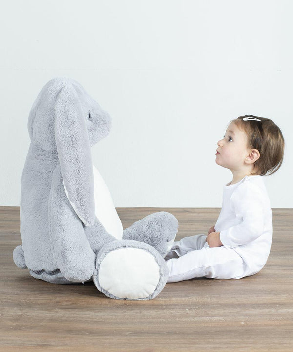 A toddler in a white outfit sits on the floor facing a large grey stuffed bunny, the MM550 by Mumbles.