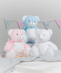 Three Mumbles MM556 teddy bears, one pink with a bow, one blue with a bow, and one white, sit on a soft blanket with pastel-colored bunting in the background.