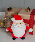A plush toy of Santa Claus is placed in front of two gift boxes wrapped in brown paper and red ribbon, with pinecones and fairy lights as decorations. The product name is MM563 from the brand Mumbles.