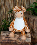 A Mumbles MM564 plush giraffe toy with a friendly expression sitting on a log, with a bamboo backdrop and green plants.