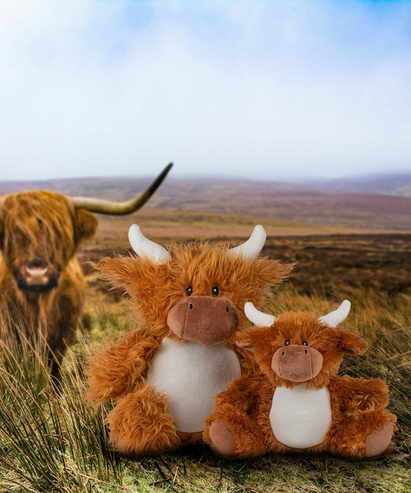 Two plush toy highland cows by Mumbles, named MM565, are positioned in grassy terrain with a real highland cow in the blurred background. The sky above is partly cloudy.