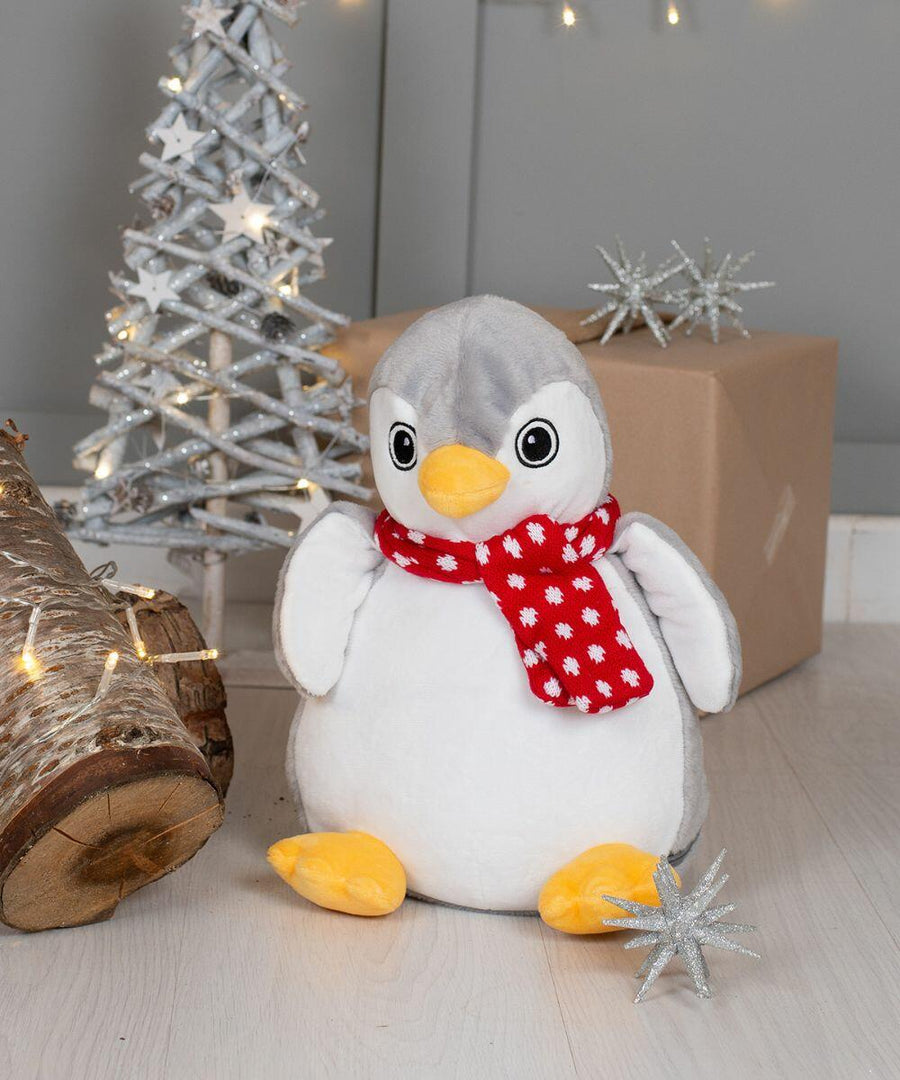 A Mumbles MM566 stuffed penguin wearing a red scarf with white snowflakes sits on a light-colored floor next to a silver decorative tree and a wrapped box in a holiday-themed setting.