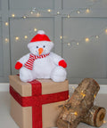 A Mumbles MM567 plush snowman toy sits on a wrapped gift box with a red ribbon. Nearby, logs adorned with string lights are placed on the floor against a gray paneled wall with hanging string lights.