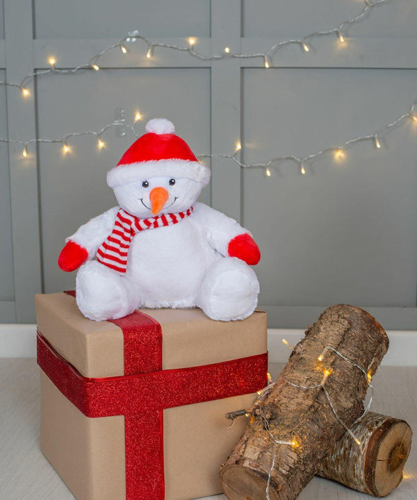 A Mumbles MM567 plush snowman toy sits on a wrapped gift box with a red ribbon. Nearby, logs adorned with string lights are placed on the floor against a gray paneled wall with hanging string lights.