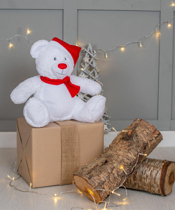 Mumbles MM573 with a red Santa hat and scarf sits on a wrapped gift box next to two logs wrapped in fairy lights. Background includes a small white tree and decorative light strings.