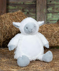 A Mumbles MM576 plush stuffed lamb toy sits against bales of hay in a rustic setting with wooden panels in the background.