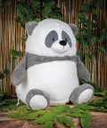 The Mumbles Zippie panda, with soft grey and white fur and an embroidered face, sits on a stone surface amid green plants, with a bamboo screen in the background.