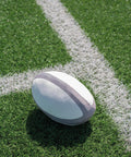 The Mumbles Zippie rugby ball, in white and grey, rests on green artificial turf near a white painted pitch line.