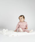 A baby in a pink outfit sits on a white blanket with an MM751 stuffed animal by Mumbles, looking towards the camera.