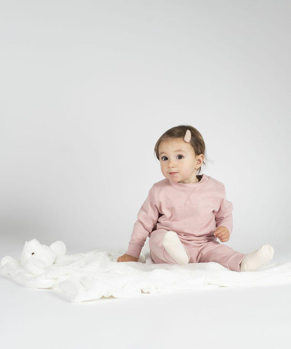 A baby in a pink outfit sits on a white blanket with an MM751 stuffed animal by Mumbles, looking towards the camera.