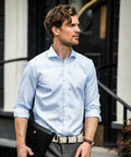 A man with wavy hair, wearing a light blue dress shirt and grey pants, holds a Nimbus N101M portfolio and stands in front of a building.