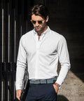 Man wearing a white shirt, dark trousers, and Nimbus N103M sunglasses, holding a black folder, walking outdoors against a dark background.