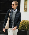 A person wearing sunglasses, a scarf, a black jacket, a grey sweater, a white shirt, and beige pants is carrying a Nimbus N104F while walking outdoors.