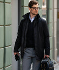 A man with glasses, wearing a black jacket over a black sweater, carries a Nimbus N105M and a pair of gloves while standing on a city street.