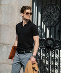 A man in sunglasses, wearing a black polo shirt and gray pants, holds a Nimbus N107M and brown folder, standing in front of an ornate iron gate.