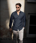 A man with Nimbus N108M sunglasses walks in partial sunlight, wearing a dark button-up shirt and light-colored pants, with his hands in his pockets.