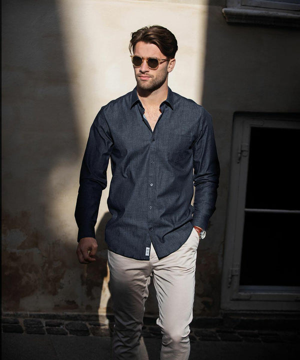 A man with Nimbus N108M sunglasses walks in partial sunlight, wearing a dark button-up shirt and light-colored pants, with his hands in his pockets.