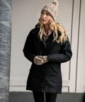 A woman with long blonde hair stands outdoors wearing a black coat, a beige Nimbus N111F knit hat with a pom-pom, and matching beige Nimbus N111F knit gloves.