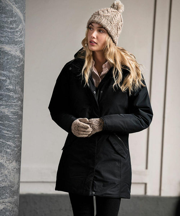 A woman with long blonde hair stands outdoors wearing a black coat, a beige Nimbus N111F knit hat with a pom-pom, and matching beige Nimbus N111F knit gloves.