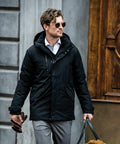 A man wearing a black coat, sunglasses, and carrying a Nimbus N111M walks past a wooden door.