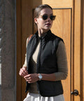 A person wearing Nimbus N114F sunglasses, a beige long-sleeve shirt, a black vest, and a white skirt stands outdoors in front of a wooden door.