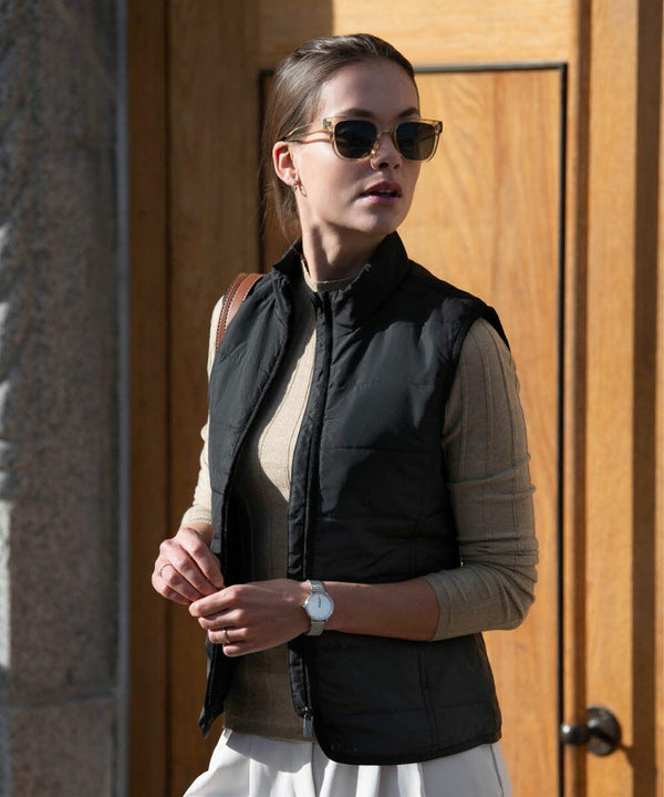 A person wearing Nimbus N114F sunglasses, a beige long-sleeve shirt, a black vest, and a white skirt stands outdoors in front of a wooden door.