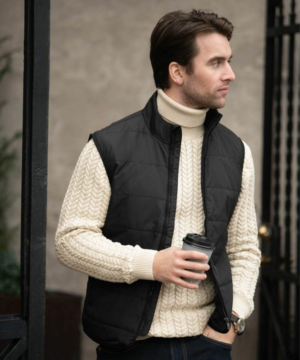 Man dressed in a cream-colored turtleneck sweater and black vest, holding a Nimbus N114M coffee cup, standing outdoors and looking to the side.