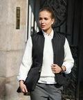 A person with blond hair, wearing a white sweater, black vest, and gray pants, holds a Nimbus N115F while walking outside in an urban setting.