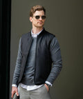 A man wearing Nimbus N115M sunglasses, a grey sweater, and a black sleeveless jacket stands in front of a dark background. He has short, wavy hair and is looking to the side.
