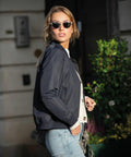A person wearing Nimbus N117F sunglasses, a navy jacket, and jeans stands outdoors near a green leafy plant, looking over their shoulder.