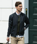 A man with short hair wearing a dark jacket, green sweater, white shirt, beige pants, and holding a Nimbus N117M bag walks in an outdoor urban setting.