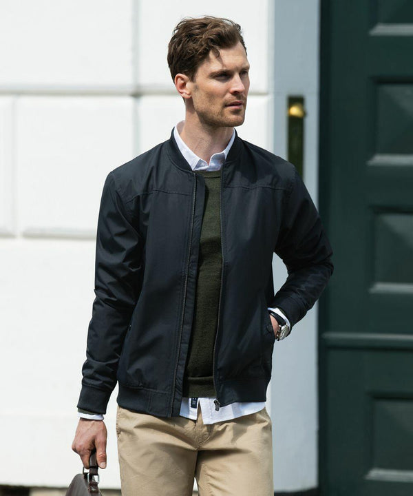 A man with short hair wearing a dark jacket, green sweater, white shirt, beige pants, and holding a Nimbus N117M bag walks in an outdoor urban setting.