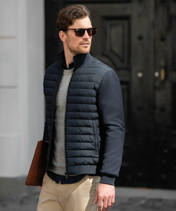 A man wearing Nimbus N122M sunglasses, a gray sweater, a black padded jacket, and beige pants holds a brown folder while standing outdoors.