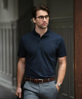 A man wearing glasses, a navy Nimbus N124M polo shirt, and gray trousers stands indoors. He has dark hair and is looking to his left.