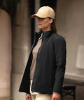 A person wearing a beige baseball cap, taupe jumper, light trousers, and a Nimbus Women's Eastlake jacket stands outdoors in an urban setting, looking to the side.