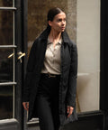 A person with dark hair pulled back is wearing the Nimbus Womens Folcroft jacket, a beige collared shirt, and black trousers while standing near a glass door with a brass handle.